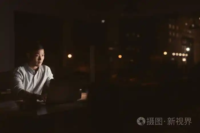 一位年轻的亚洲商人深夜独自在办公室的办公桌前加班,背景是城市的