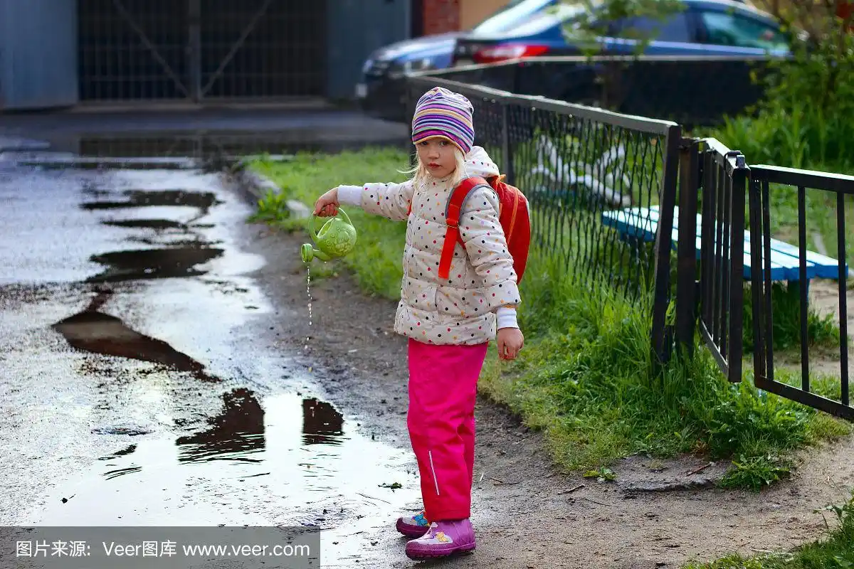 蹒跚学步的小女孩穿着五颜六色的雨衣和雨靴在泥泞的水坑里用喷壶浇水