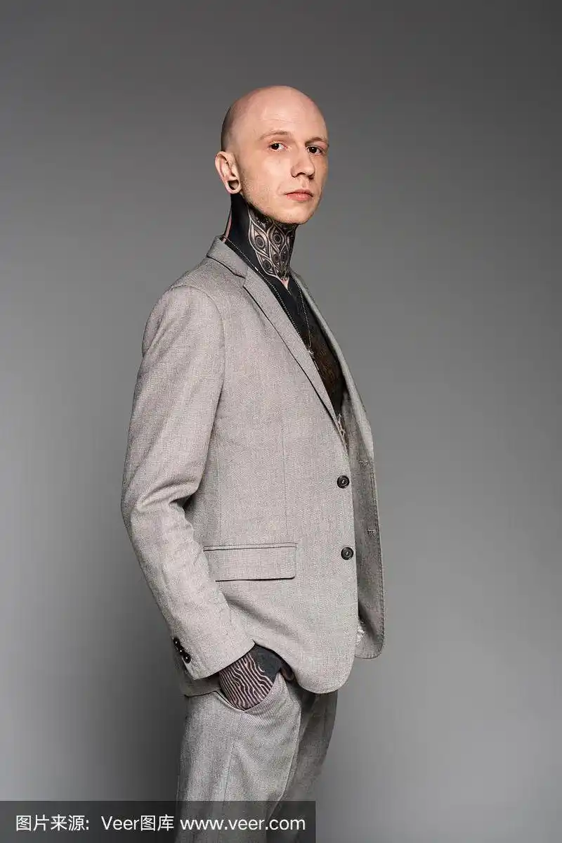 侧面的英俊的秃头纹身男子在西装站在口袋和看孤立的灰色相机