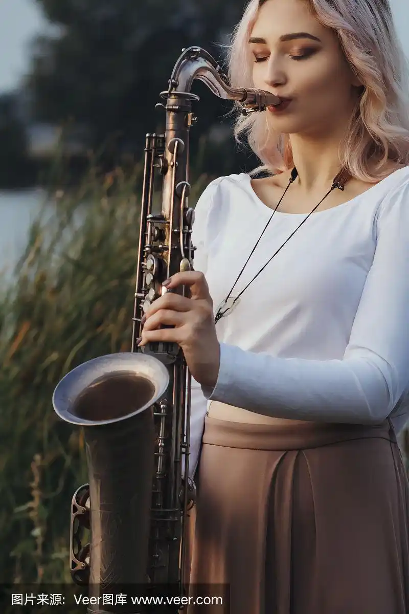 年轻女子在河边吹萨克斯风的肖像,金发女孩与乐器在户外的日出,概念