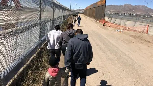 migrants trudge along the border fence to a waiting bus after